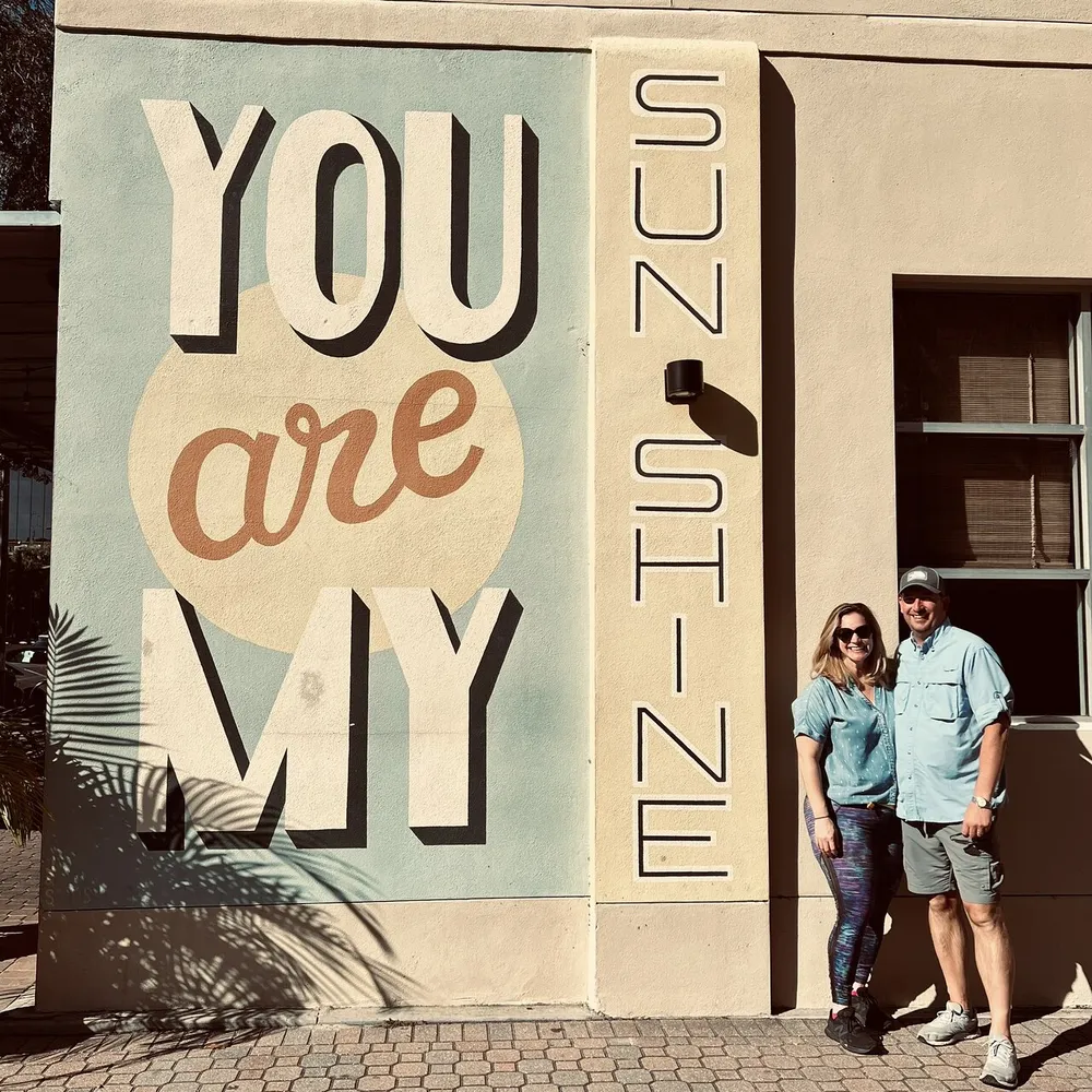 A couple is standing in front of a wall with a large mural that reads YOU ARE MY SUNSHINE in stylized letters