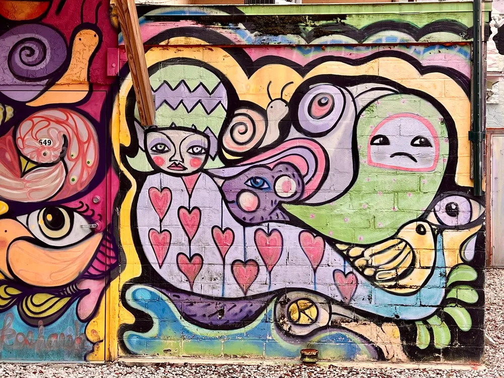 The image shows a colorful whimsical mural featuring an assortment of stylized creatures and patterns including a figure with a crown hearts and eyes with a vibrant street art aesthetic