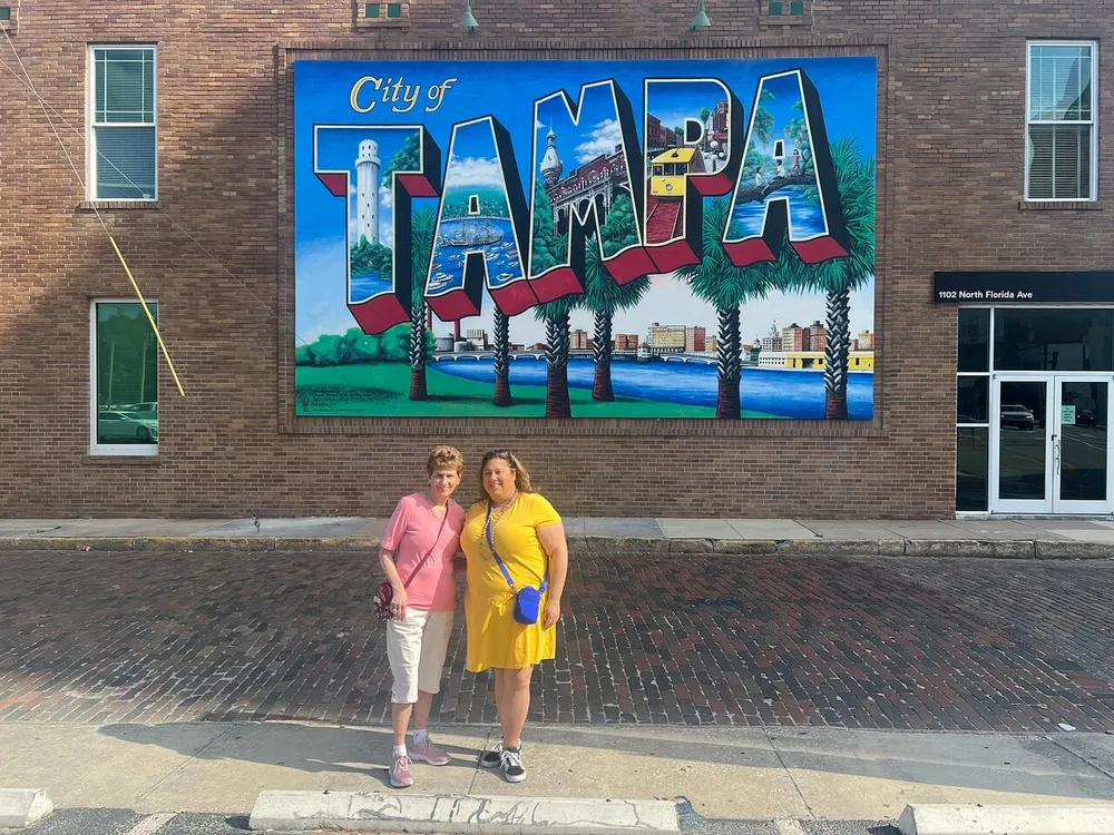 Two people are posing for a photo in front of a vibrant mural that spells City of Tampa