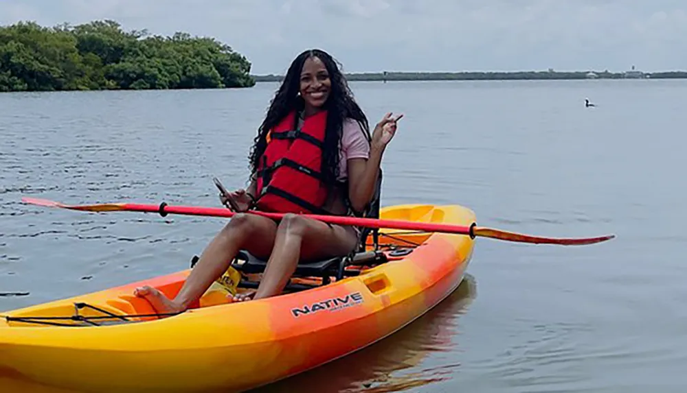 A smiling person is sitting in a bright yellow and orange kayak on calm water holding a paddle and flashing a peace sign