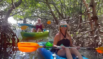 Two people are kayaking through a mangrove forest, with the person in the foreground smiling at the camera.