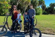 A family of four is posing with two bicycles on a sunny day in a park with palm trees in the background.