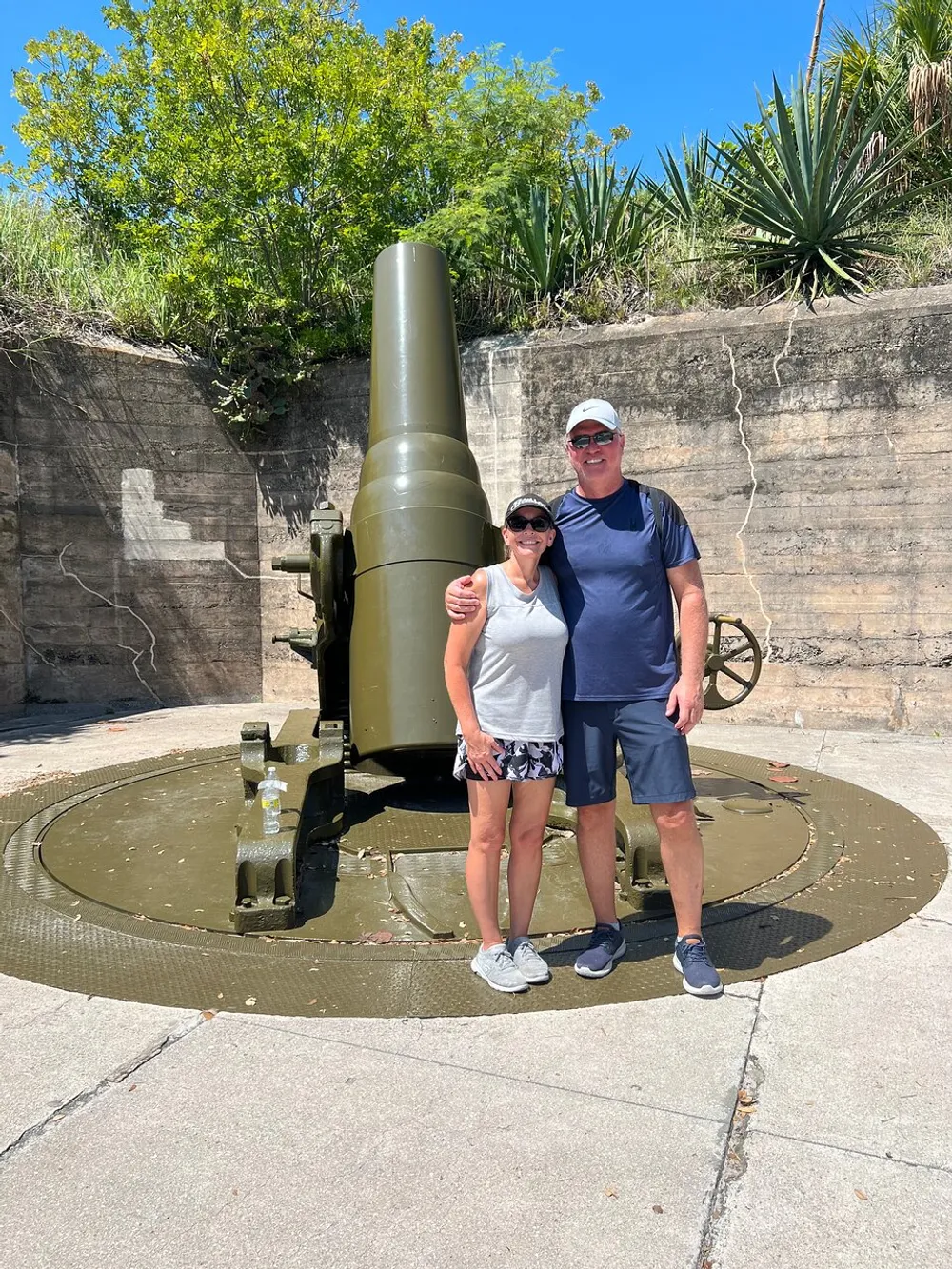 Two people are smiling for a photo in front of a large historic cannon on a sunny day