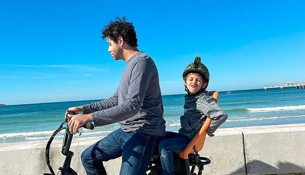 A man and a child are enjoying a bike ride along a beachside path under a clear blue sky