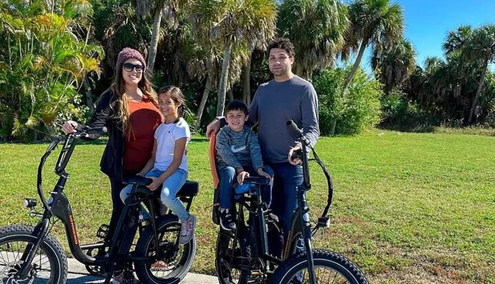 A family of four is standing outdoors with two electric bikes smiling for the camera with a backdrop of palm trees and greenery