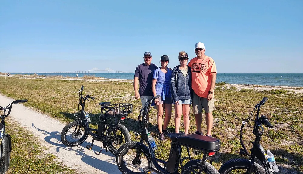 Four people are standing by the seaside with bicycles in the foreground and a bridge is visible in the background under a clear sky