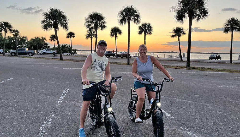 Two people are smiling and posing on their bicycles during sunset by a palm-lined waterfront