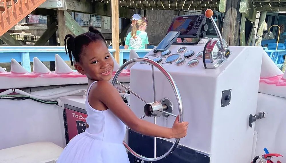 A smiling young girl is standing at the helm of a boat pretending to steer with the wheel