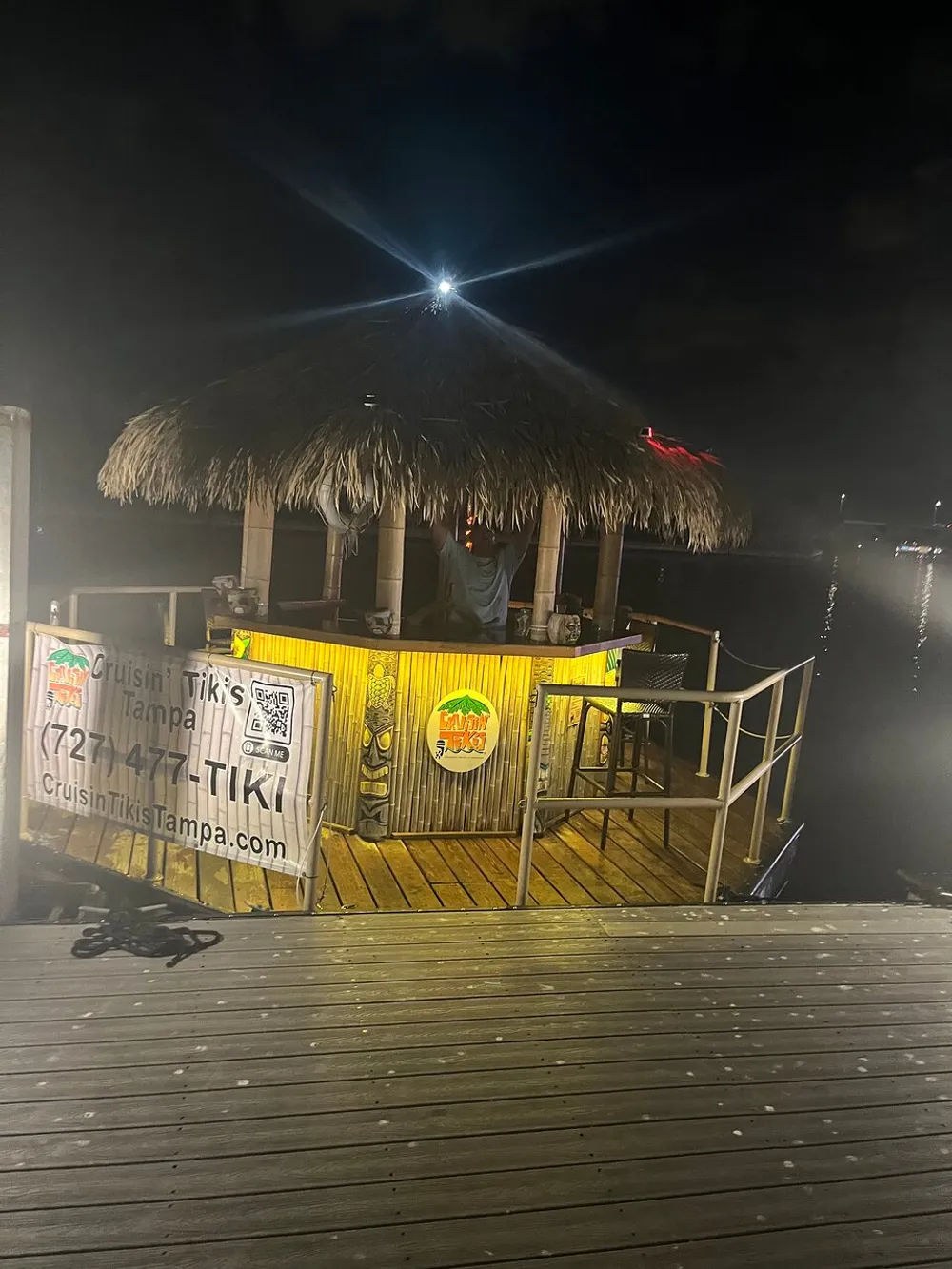 The image shows a thatch-roofed tiki bar by a waterfront at night illuminated with a bright light at the top and signage promoting a cruising tiki experience
