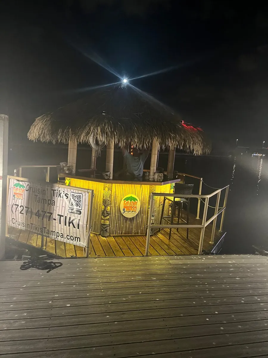 The image shows a thatch-roofed tiki bar by a waterfront at night, illuminated with a bright light at the top and signage promoting a cruising tiki experience.