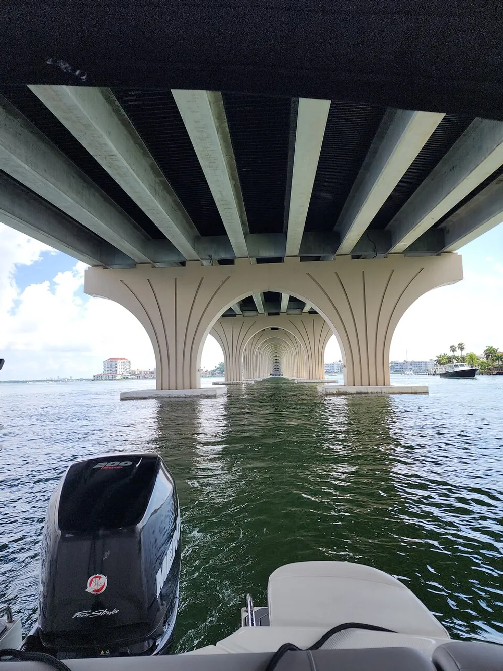 A boat is poised to pass under a long arched concrete bridge spanning a body of water creating a symmetrical tunnel-like effect