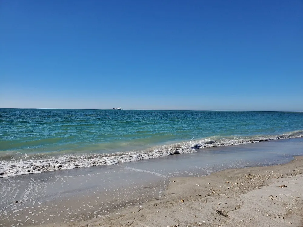 A serene beach scene with gentle waves lapping on the shore under a clear blue sky with a distant ship on the horizon