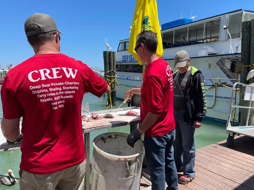 Two crew members in red shirts are working on a dock next to a boat handling fish with a bystander observing
