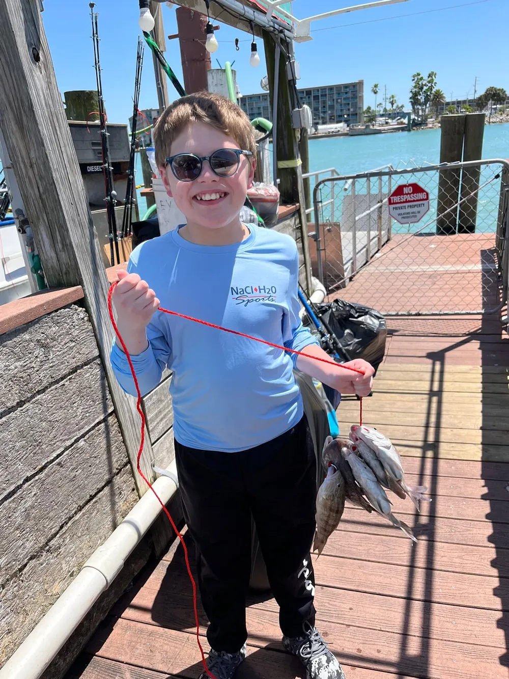 A smiling young boy wearing sunglasses is proudly holding up a string of fish possibly showcasing his catch from a day of fishing at a dock