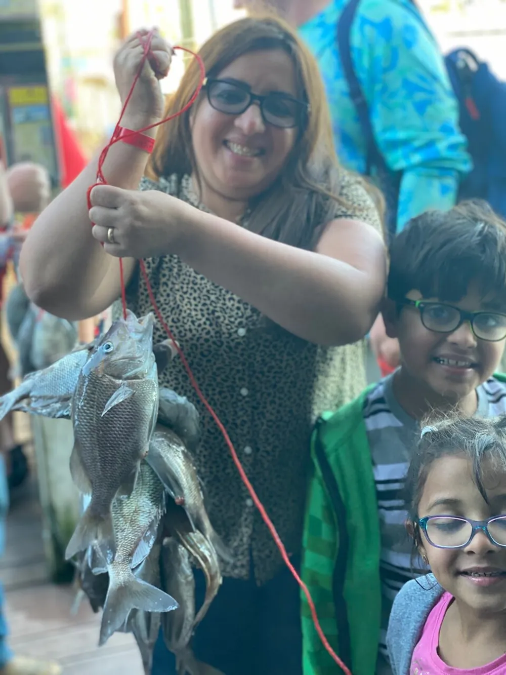 A smiling woman proudly displays several fish she appears to have caught with two cheerful kids looking on