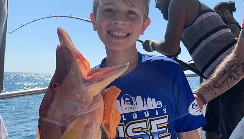 A smiling young boy is posing on a boat proudly displaying a large colorful fish he seemingly caught with others and fishing equipment visible in the background