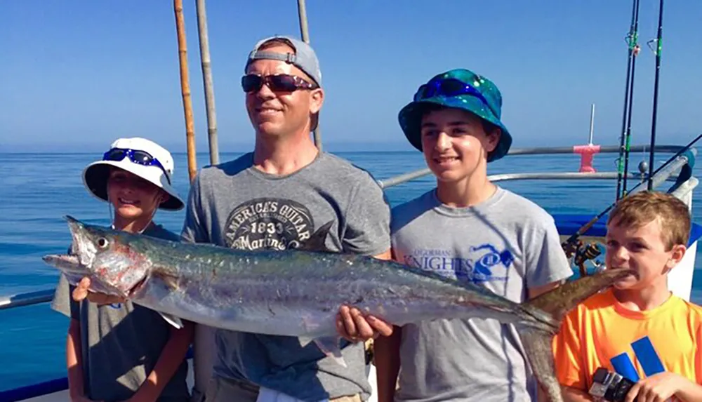 A group of four people likely a family are posing on a boat with fishing rods in the background while one of them holds a large fish they presumably just caught