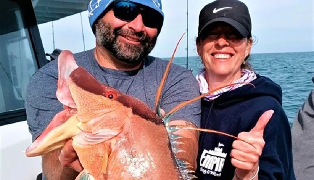A man and a woman are smiling on a boat proudly displaying a large reddish fish they have caught
