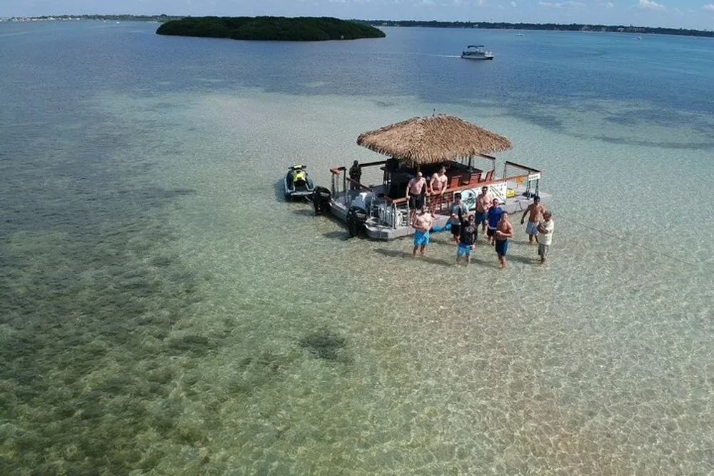 A group of people is standing in shallow clear waters near a floating tiki bar structure with a boat tied to it with islands visible in the background