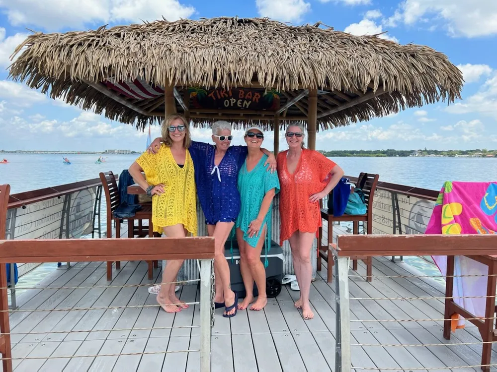 Four smiling individuals are standing in front of a tiki bar with a thatched roof on a wooden deck by the water with bright towels and clear skies contributing to a leisurely tropical atmosphere