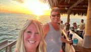 A smiling couple takes a selfie at a seaside bar with a beautiful sunset in the background.