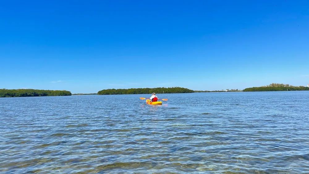 A person kayaks on tranquil blue waters with green islands in the distance under a clear sky