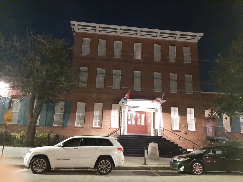 The image shows a nighttime view of a red-brick multi-story building with American and state flags above the entrance flanked by parked cars under street lighting
