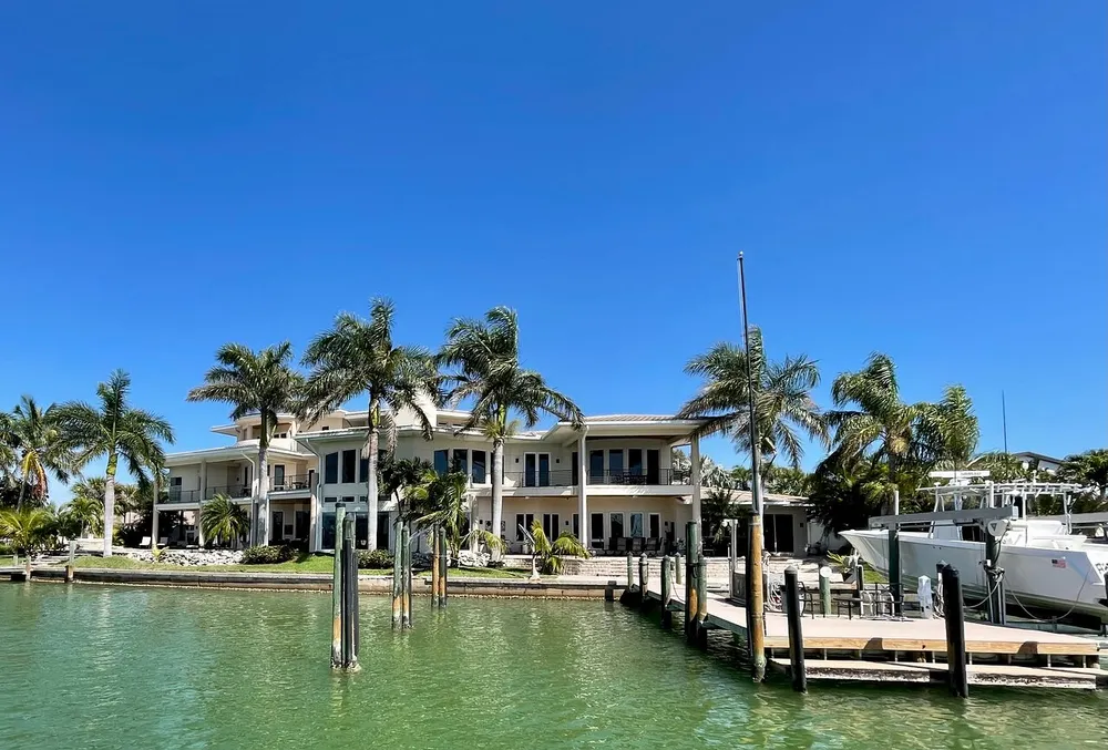 The image shows a luxurious waterfront house with a clear blue sky surrounded by palm trees and a boat docked at a private pier