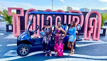 Three individuals are posing for a photo in front of large pink letters spelling 