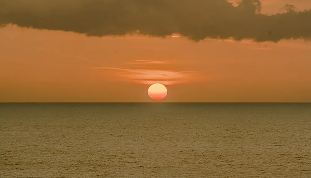 The image captures a serene sunset with a large glowing sun poised just above the horizon of a tranquil sea under a sky with hues of orange and a hint of cloud cover