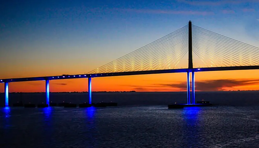 A bridge illuminated with blue lights stands against a dramatic sunset sky reflected in the water below