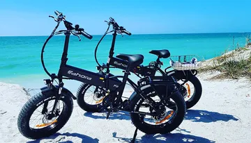 Two black electric fat-tire bicycles are parked on a sandy beach with a clear blue sea in the background.