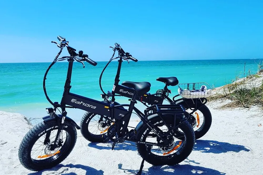 Two electric bicycles are parked on a sandy beach against a backdrop of vibrant blue sea and sky