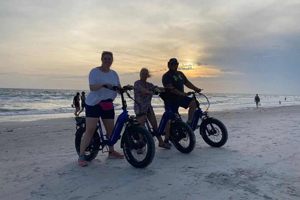 Three people are smiling for the camera while standing with their bicycles on a sandy beach at dusk