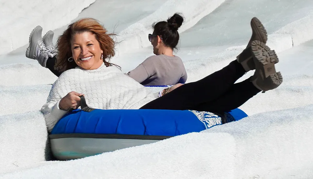 Two individuals are enjoying a sunny day on a snowy hill with the person in the foreground smiling broadly as they slide down on a blue inflatable snow tube