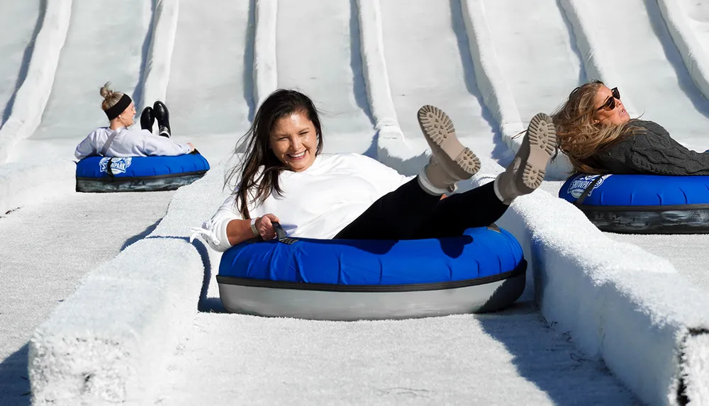 Three people are gleefully sliding down a snowy hill on blue inflatable inner tubes