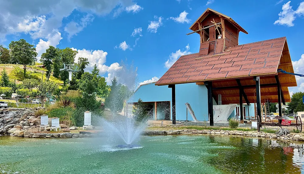 The image shows a scenic view of a pond with a fountain in front of a building under construction with a lush green hill and ski lift chairs in the background under a blue sky dotted with clouds