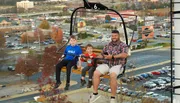 A man and two children are riding on a chairlift with a cityscape in the background.