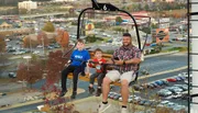 A man and two children are riding on a chairlift with a cityscape in the background.