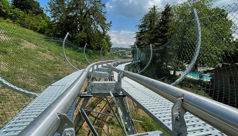 A metal summer toboggan slide meanders down a green hillside with safety nets on either side under a partly cloudy sky