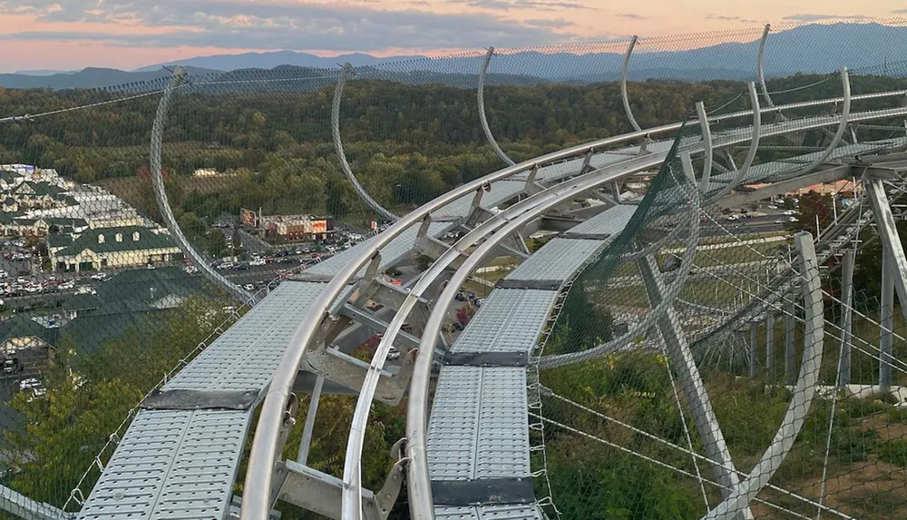 This image shows a curving metal slide structure of an alpine coaster winding down a hill with a view of a landscape with buildings and distant mountains at twilight