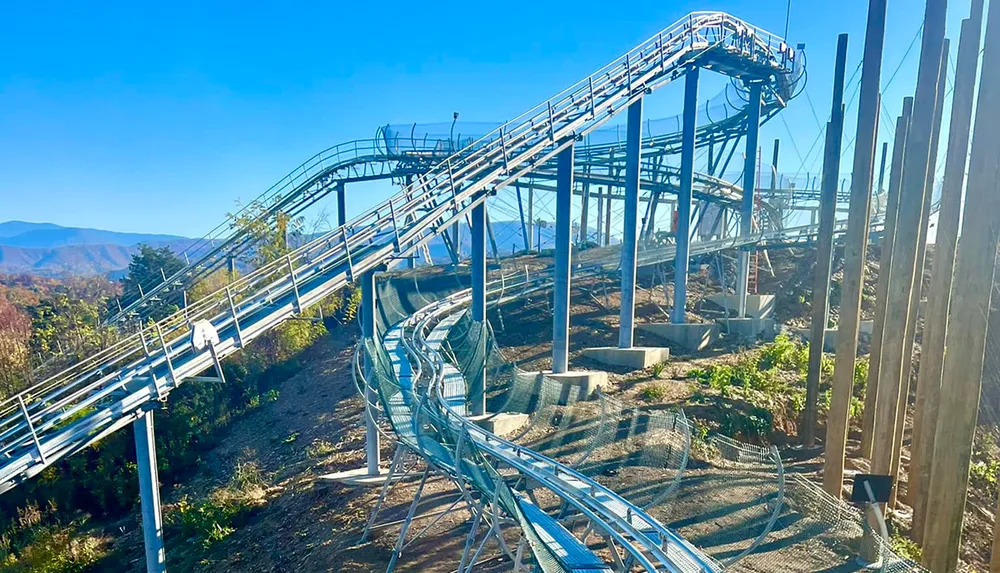 The image shows an empty mountain coaster winding through a hilly tree-dotted landscape under a clear blue sky