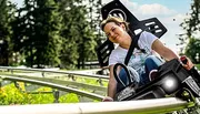 A person is enjoying a ride on an alpine coaster amidst green surroundings on a sunny day.