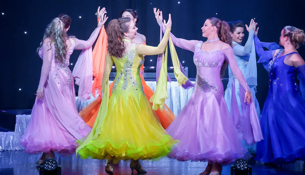 This image shows a group of performers in colorful sparkling dresses dancing on stage with an air of joy and celebration