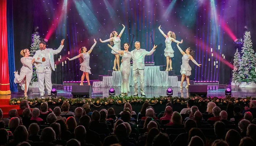 A group of performers dressed in sparkling outfits dance on a stage decorated with festive lights and ornaments, in front of an audience.