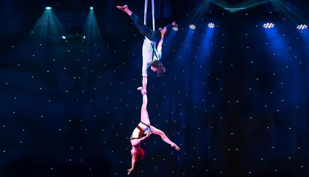 Two aerial performers executing a dramatic and elegant hanging pose on stage with one suspended from above and the other hanging upside down