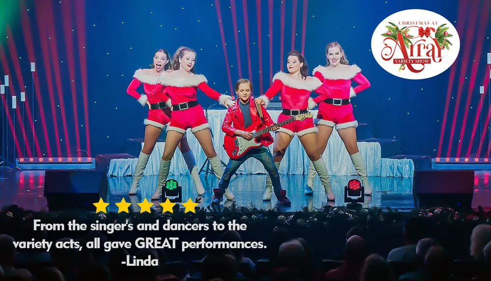 A young boy in a red jacket plays an electric guitar on stage surrounded by four dancers in festive red and white outfits