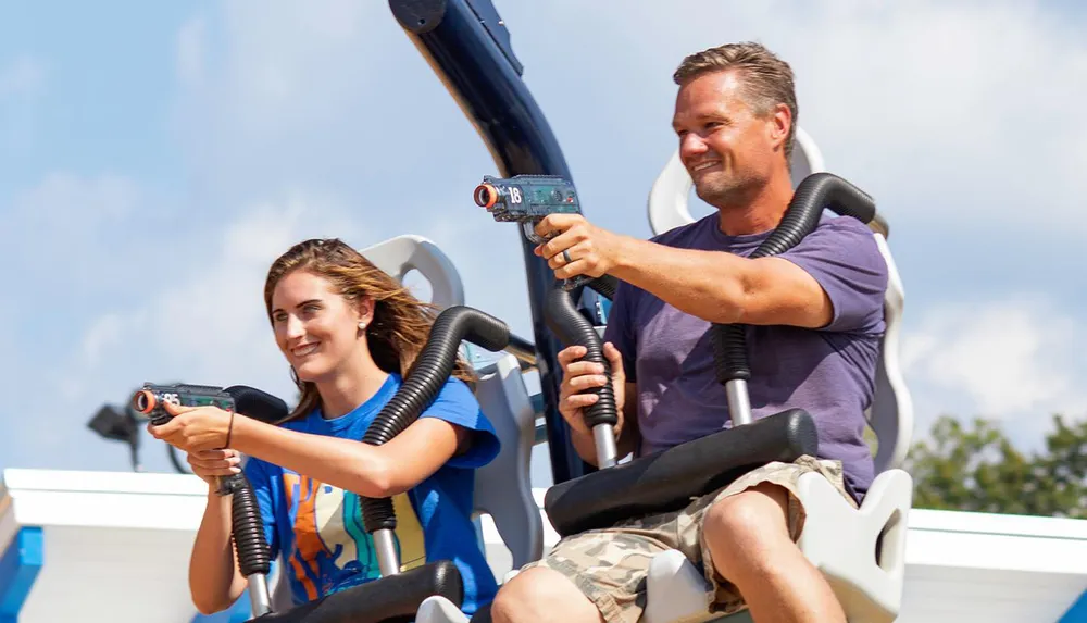 Two people are enjoying a ride at an amusement park smiling while holding controls that may be part of an interactive game