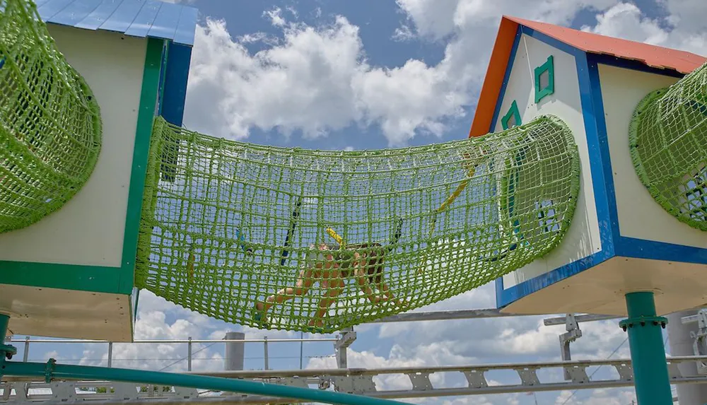 A child is traversing a green rope bridge at a colorful playground under a blue sky dotted with clouds