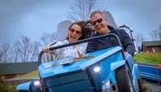 Two people are joyfully experiencing a ride in a blue roller coaster car, with dynamic expressions and windswept hair.